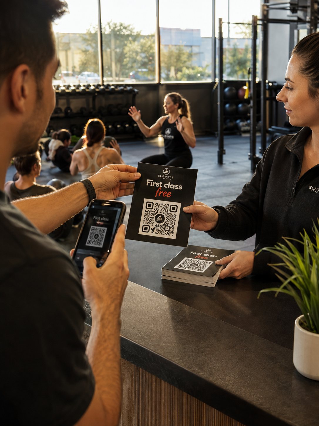 A gym front desk handing a new visitor a First Class Free flyer while he scans its code, the coach mid-class behind